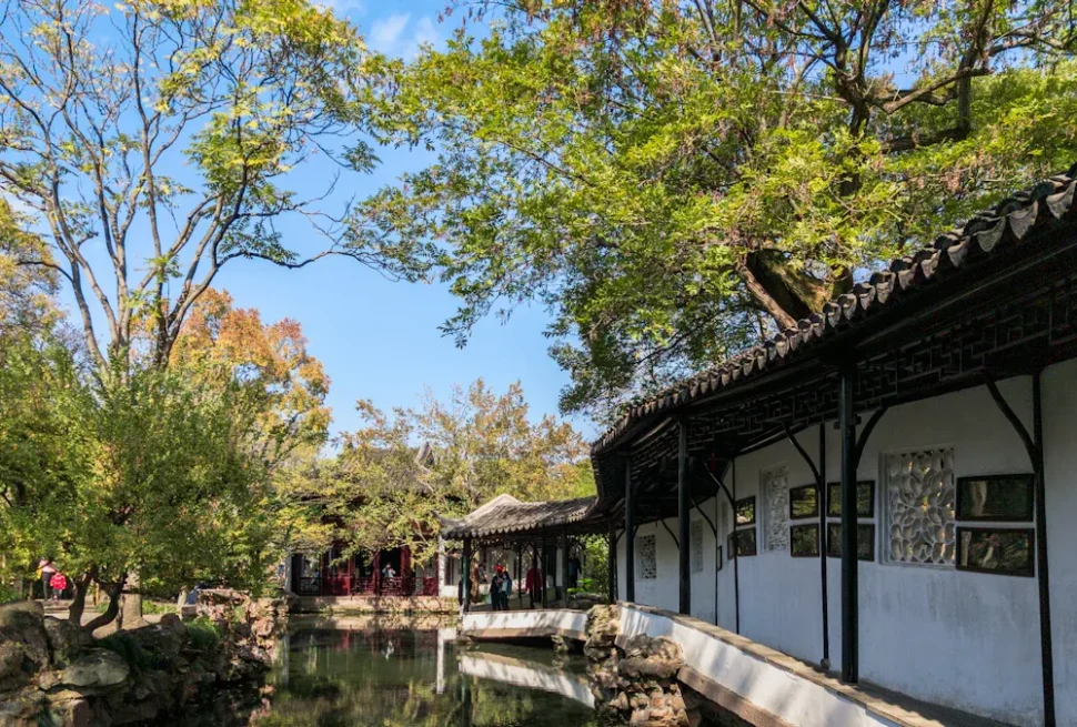 Traditional Chinese garden with white-walled buildings, dark wooden beams, curved-tile roofs, and a reflecting pond.
