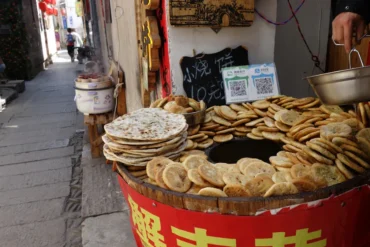 Street food stall with a circular pan surrounded by dozens of flatbreads in a busy alley.