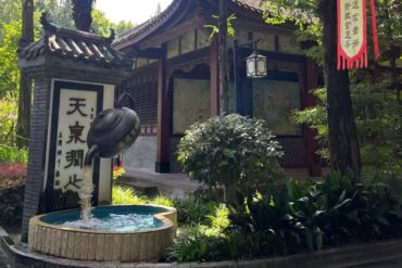 Teapot-shaped fountain pouring water into a round basin beside a traditional Chinese garden pavilion.