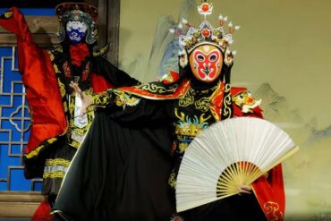 Two performers in ornate Chinese opera costumes with painted masks; one holds a white fan while the other strikes a pose in a red-and-black outfit.
