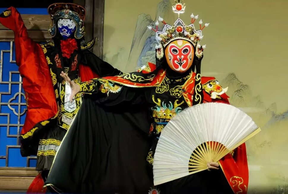 Sichuan Opera Face Changing,Chengdu,Chinatour Two performers in ornate Chinese opera costumes with painted masks; one holds a white fan while the other strikes a pose in a red-and-black outfit.