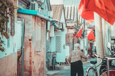 Older woman walks down a narrow alley between colorful old buildings with red Chinese flags overhead.