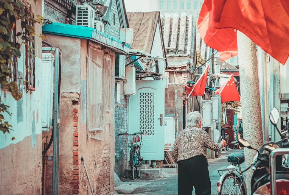 Older woman walks down a narrow alley between colorful old buildings with red Chinese flags overhead.