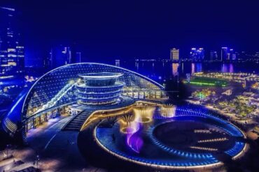 Hangzhou Grand Theatre Shows ,Hangzhou,Chinatour Nighttime aerial view of a futuristic blue-lit urban complex with a curved bridge, circular plaza, and fountains near the water.