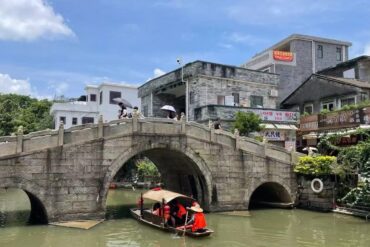 Shiwan Ancient Towns,Foshan,Chinatour Stone arch bridge over a canal with pedestrians holding umbrellas on the bridge and a small tour boat passing beneath.