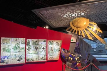 Museum exhibit with a red wall displaying comic panels and Chinese characters alongside a large metallic sunburst sculpture roped off in the foreground.