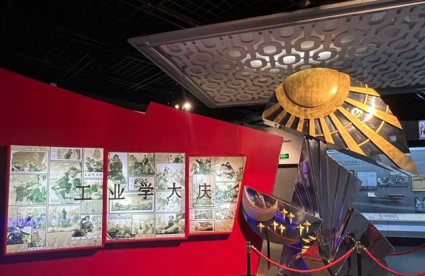 Museum exhibit with a red wall displaying comic panels and Chinese characters alongside a large metallic sunburst sculpture roped off in the foreground.