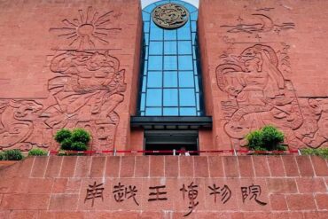 Front view of a red brick museum facade featuring large stone reliefs on both sides, a blue glass central tower, and Chinese characters along the lower wall.