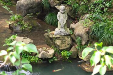 Lingyin Temple ,Hangzhou,Chinatour Stone statue of a small monk standing on a rock beside a koi pond, surrounded by green plants and rocks in a tranquil garden scene with a swimming fish nearby.