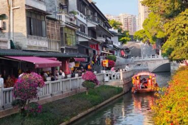 Lychee Bay Water Town,Guangzhou,Chinatour Crowded riverside street with shops and pink flowers along a canal, a colorful boat gliding by.