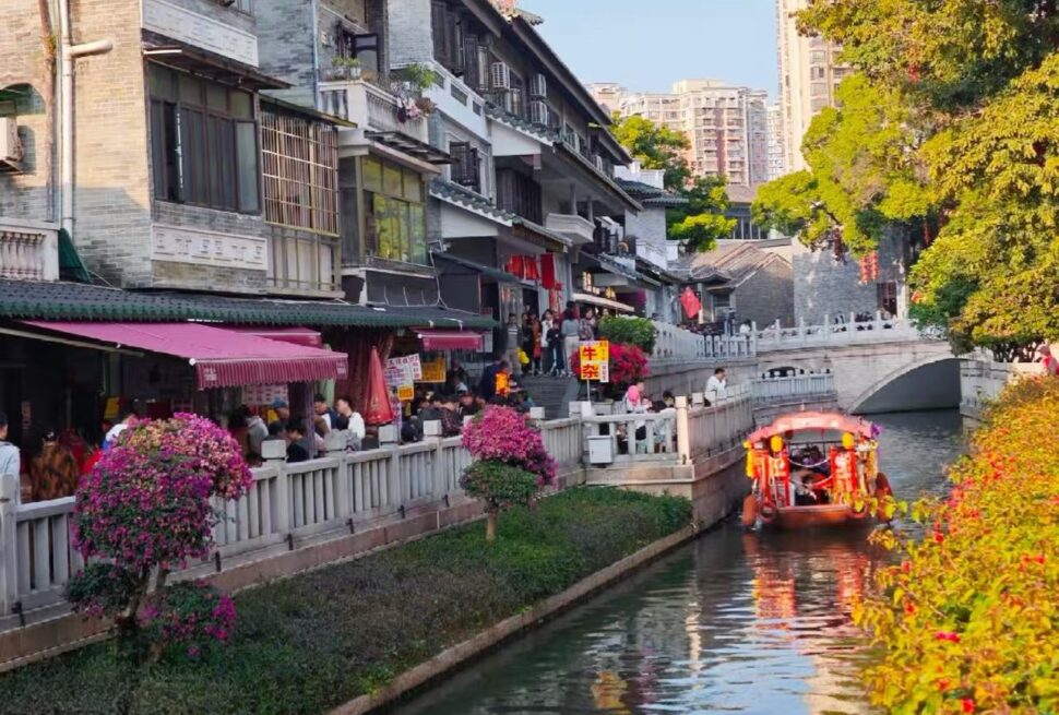 Crowded riverside street with shops and pink flowers along a canal, a colorful boat gliding by.