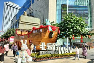 Large wooden ship sculpture decorated with red ribbons sits in a busy city square, with a colorful bunny statue and a big foreground sign reading “Walk” near shoppers and modern buildings.