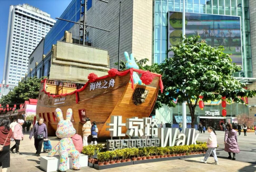 Large wooden ship sculpture decorated with red ribbons sits in a busy city square, with a colorful bunny statue and a big foreground sign reading “Walk” near shoppers and modern buildings.