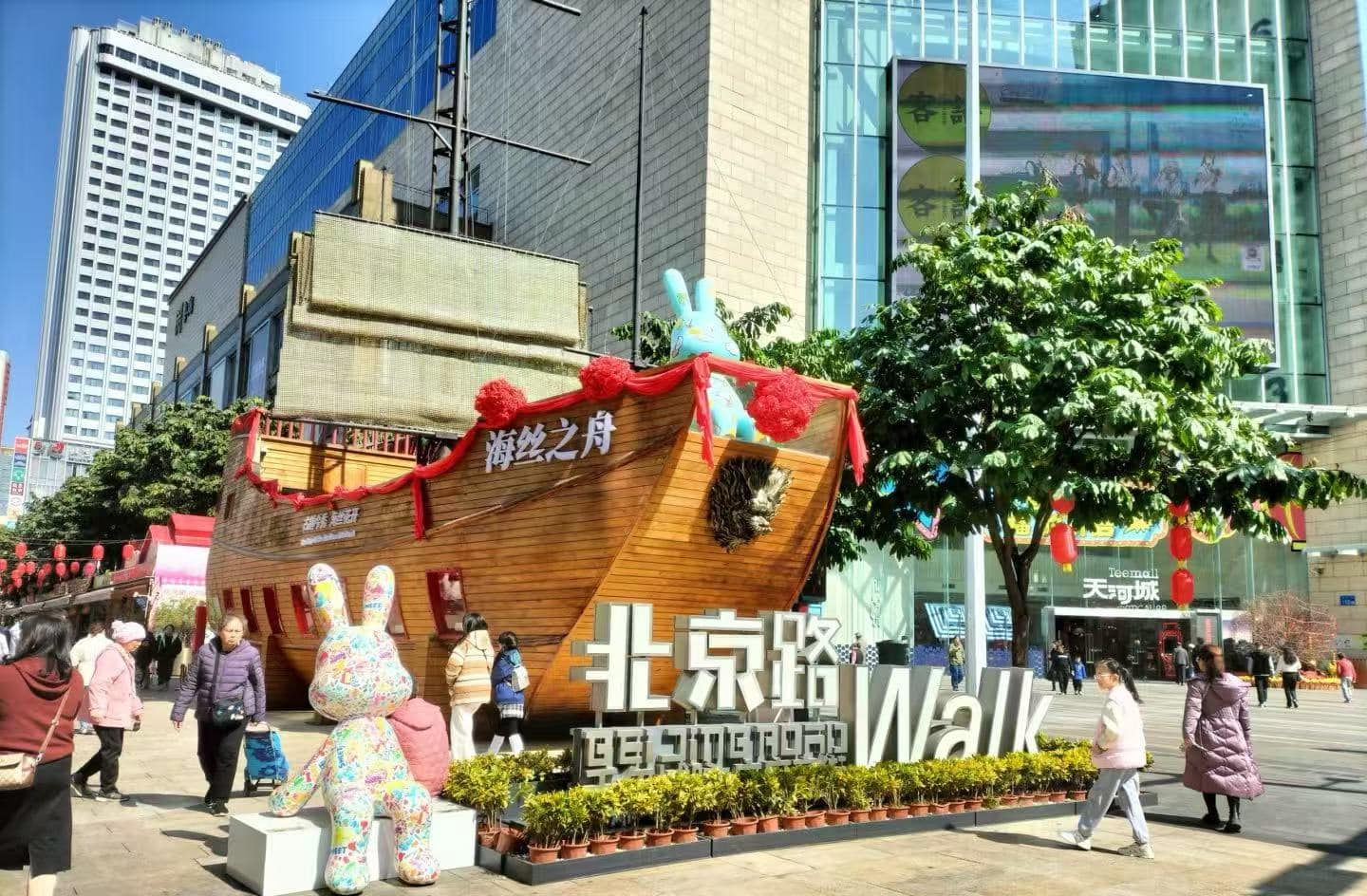 Beijing Road,Guangzhou,Chinatour Large wooden ship sculpture decorated with red ribbons sits in a busy city square, with a colorful bunny statue and a big foreground sign reading “Walk” near shoppers and modern buildings.