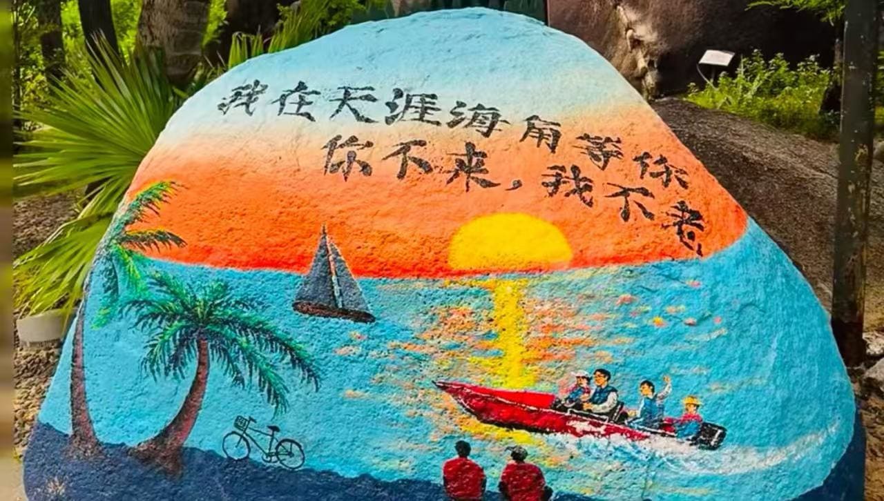 Colorful mural-painted rock showing sunset over the sea, palm trees, a sailboat, a red motorboat with people, two onlookers, a bicycle silhouette, and Chinese text above.