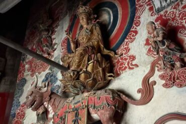 Golden crowned deity seated on a dragon, mounted on a richly painted temple wall backdrop of red cloud motifs.