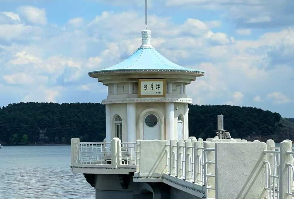 White round pavilion with a blue-teal roof on a pier over calm water, with trees in the distance.