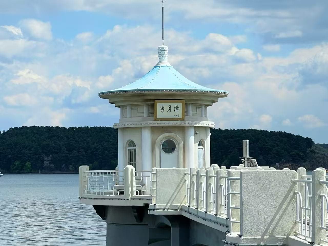 Jingyuetan National Forest Park,Changchun,Chinatour White round pavilion with a blue-teal roof on a pier over calm water, with trees in the distance.