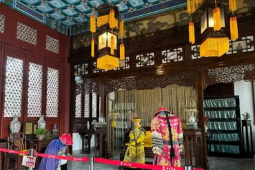 New Yuanming Palace,Zhuhai,Chinatour Inside a traditional Chinese hall with dark carved wood, gold-yellow lanterns, and red walls; two people in ornate dynastic costumes stand near a ceremonial altar.