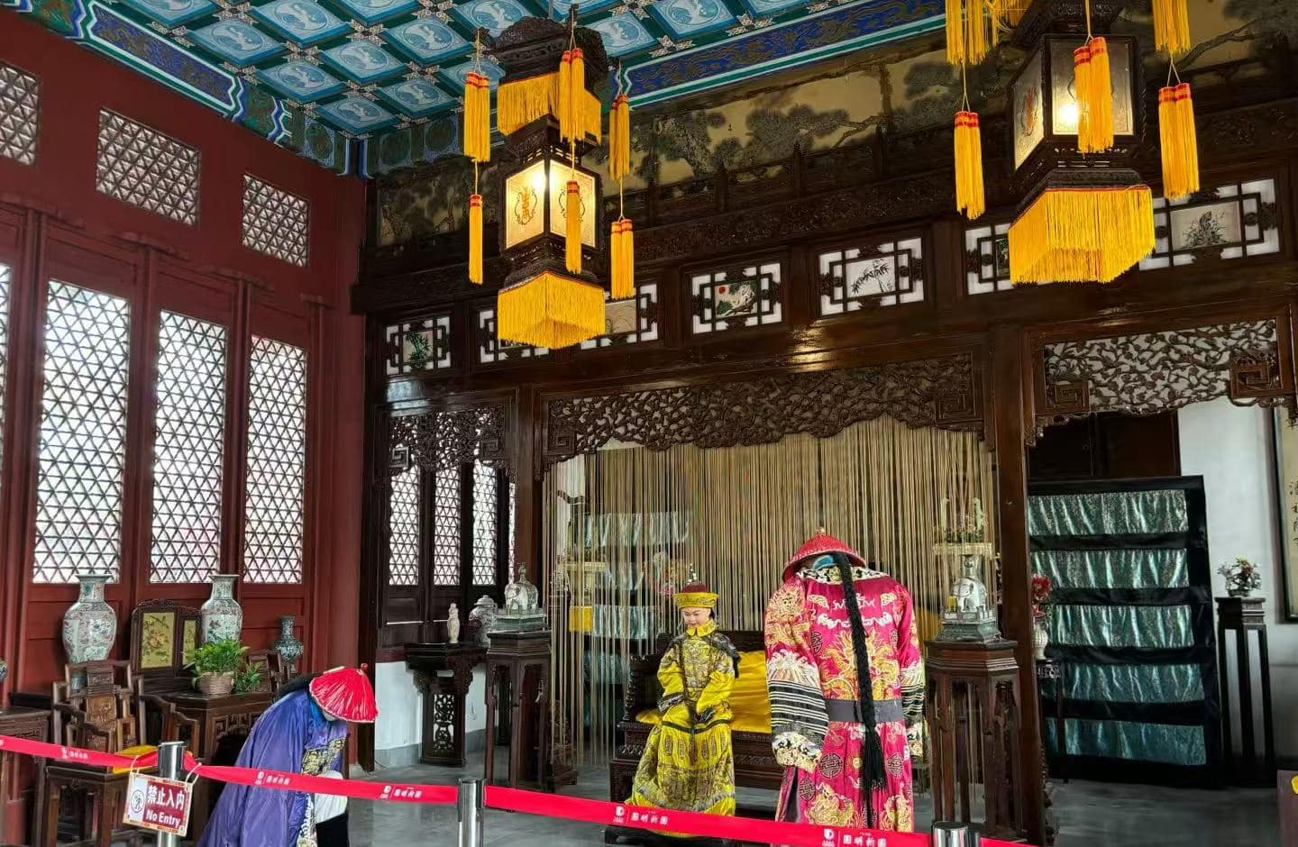 New Yuanming Palace,Zhuhai,Chinatour Inside a traditional Chinese hall with dark carved wood, gold-yellow lanterns, and red walls; two people in ornate dynastic costumes stand near a ceremonial altar.