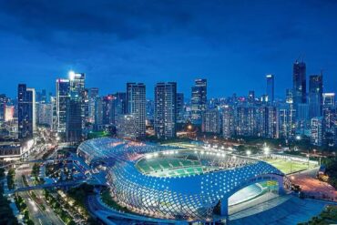 Aerial night view of a futuristic stadium with a blue lattice roof beside a dense illuminated city skyline.