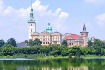 European-style castle complex with tall spires by a tree-lined riverfront, reflection in the water