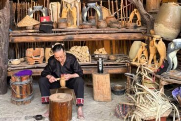 Betel Nut Valley Binglanggu Li and Miao Culture,Sanya,Chinatour An elderly artisan carving a wooden figure in a rustic workshop filled with pottery and wooden sculptures on shelves.