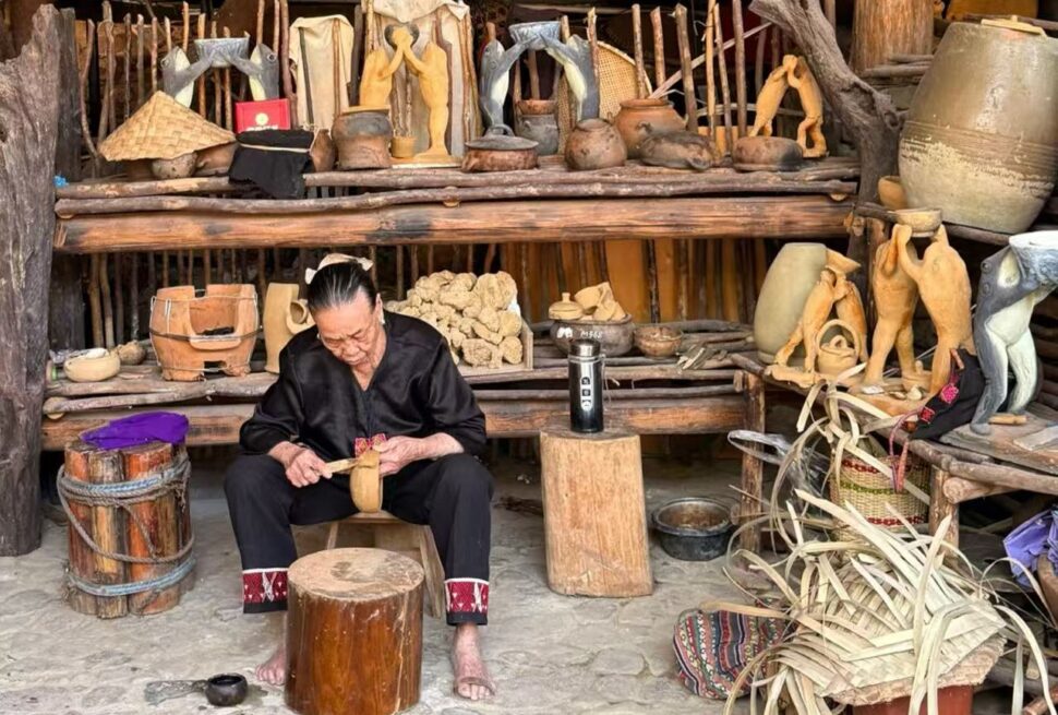 Betel Nut Valley Binglanggu Li and Miao Culture,Sanya,Chinatour An elderly artisan carving a wooden figure in a rustic workshop filled with pottery and wooden sculptures on shelves.