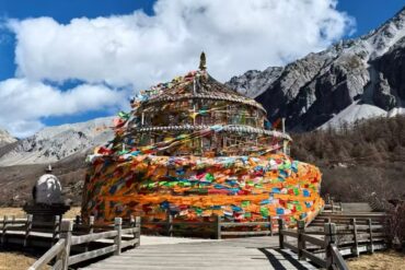Colorful multi-tiered prayer wheel surrounded by wooden walkway against a rocky mountain landscape and bright blue sky with clouds
