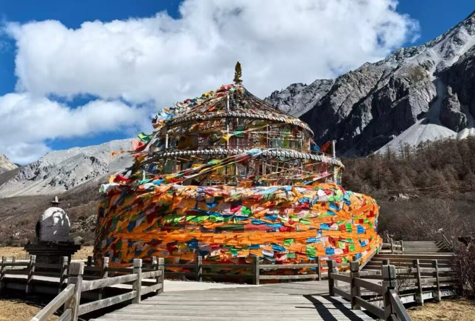 Colorful multi-tiered prayer wheel surrounded by wooden walkway against a rocky mountain landscape and bright blue sky with clouds