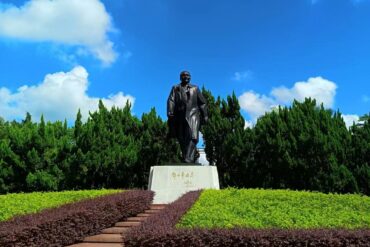 Bronze statue of a man in a suit standing on a pedestal in a landscaped park with shrubs and steps.
