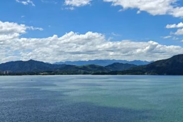 Taiping Lake,Huangshan,Chinatour Calm turquoise lake with distant green mountains under a bright blue sky and fluffy white clouds; a small town sits along the shoreline on the left.