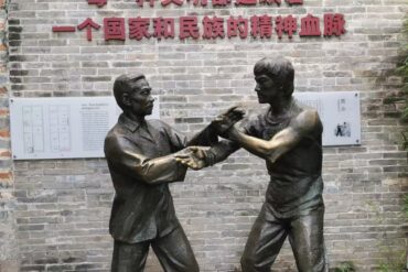Bronze statue of two men sparring in martial arts in front of a brick wall with Chinese inscription.