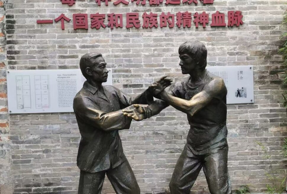 Bruce Lee’s Ancestral Home,Foshan,Chinatour Bronze statue of two men sparring in martial arts in front of a brick wall with Chinese inscription.