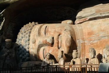 Dazu Rock Carvings Buddhist Sites,Chongqing,Chinatour Large reclining Buddha carved in rock inside a cave, with several standing and seated stone figures nearby and a railing in front.