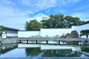 Suzhou Museum,Suzhou,Chinatour Serene Chinese garden with white walls, a reflective pond, rocky rock garden, and a glass pyramid sculpture reflected on the water under a blue sky.