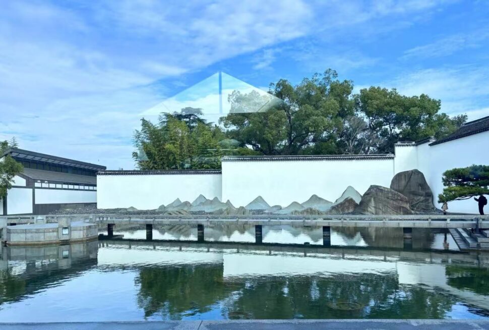 Serene Chinese garden with white walls, a reflective pond, rocky rock garden, and a glass pyramid sculpture reflected on the water under a blue sky.