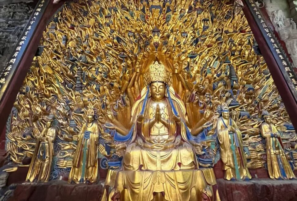 Golden statue of a multi-armed bodhisattva seated in meditation, surrounded by many outstretched arms and smaller figures in a temple setting.