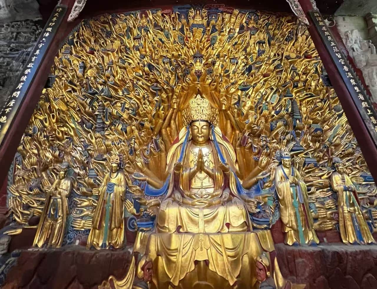 Golden statue of a multi-armed bodhisattva seated in meditation, surrounded by many outstretched arms and smaller figures in a temple setting.