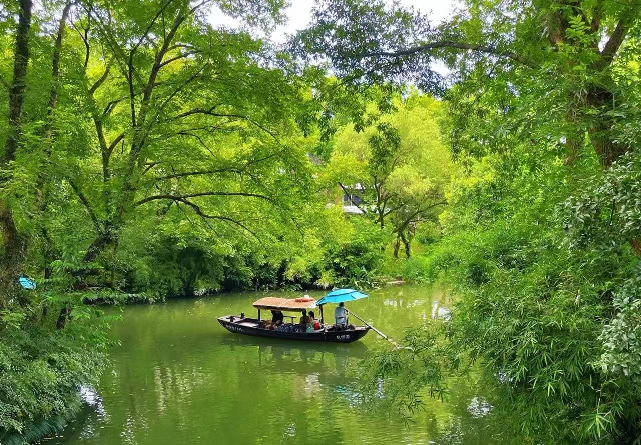 A wooden boat with a blue umbrella carries passengers along a calm, green river surrounded by lush trees and foliage.