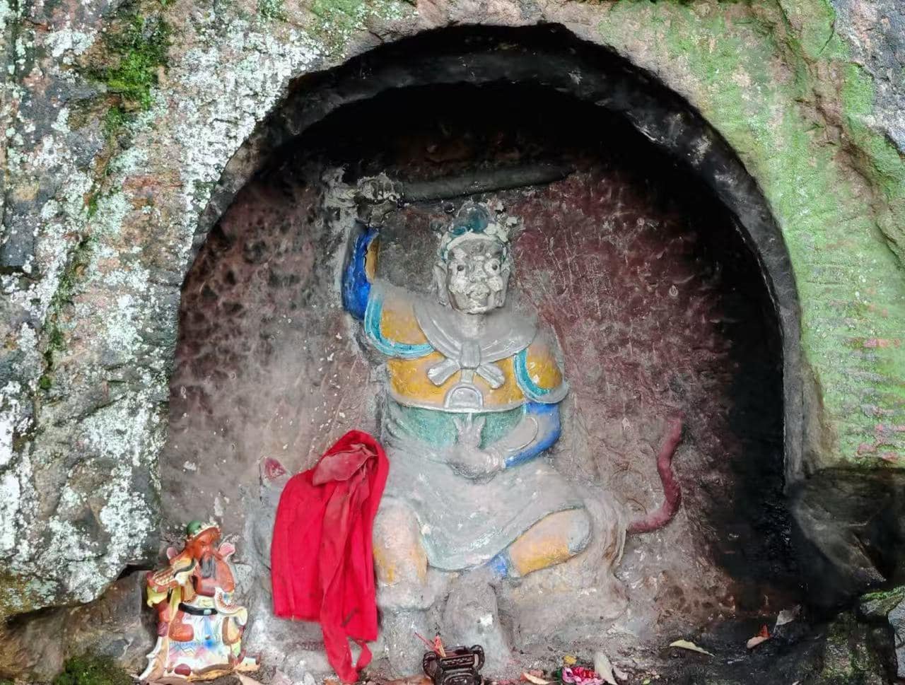 Statue of a seated deity carved into a rocky cave, with yellow and blue clothing, a red cloth draped nearby, and small figurines around the altar in a moss-covered alcove.