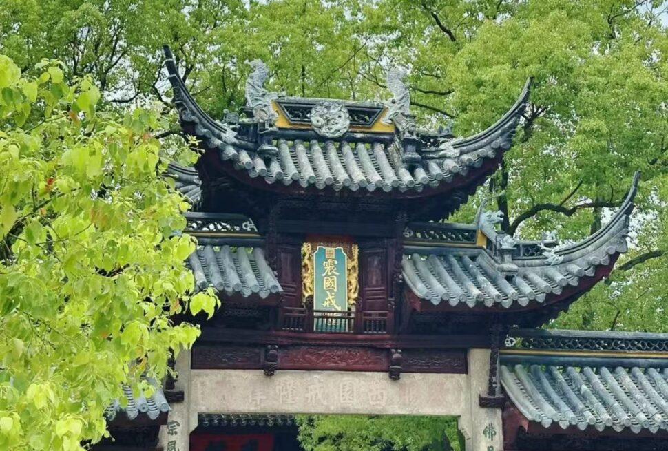 Traditional Chinese temple gate with curved eaves and ornate roof, green signboard with gold characters, surrounded by trees.