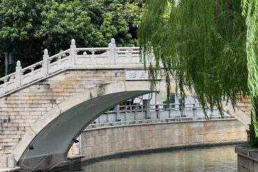 Stone arched bridge over a narrow canal, with a sloped stone ramp and hanging willow branches on the right; lush trees in the background.