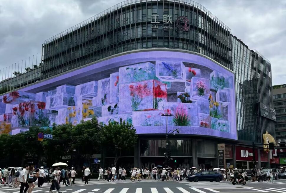 Busy city street scene with a curved LED screen on a glass-fronted building displaying colorful flower images as pedestrians cross the crosswalk.