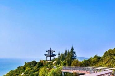 Traditional pagoda on a hillside overlooking a blue sea, with a curved wooden boardwalk in the foreground.