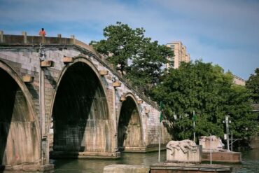 Grand Canal,Suzhou,Chinatour Stone arched bridge over a river with green trees nearby; a person in an orange shirt stands on the walkway above.