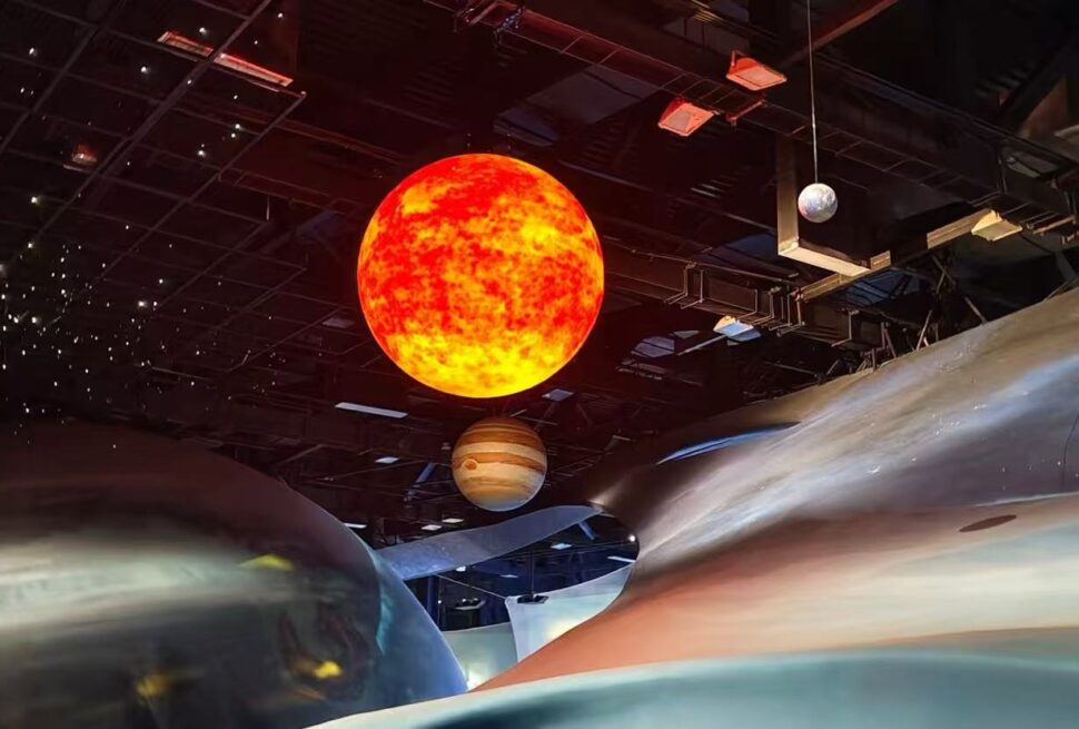 Large bright orange sun model suspended from the ceiling with a Saturn-like planet nearby in a dark museum exhibit.