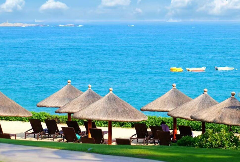 Tropical beach with thatched umbrellas and lounge chairs facing a blue sea; small boats drift on calm water.
