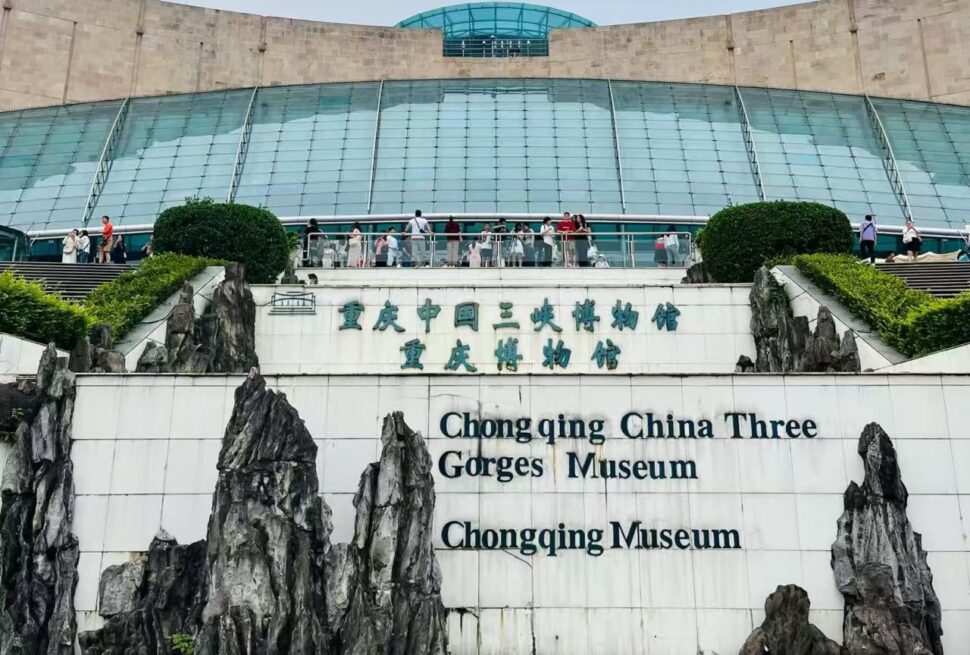Exterior view of Chongqing China Three Gorges Museum with a glass-domed atrium, visitors on the terrace, and rock sculptures framing a sign that reads 'Chongqing China Three Gorges Museum' in English and Chinese.