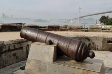 Humen Fort,Dongguan,Chinatour Antique rusted cannon mounted on a stone pedestal at a coastal fort, with a misty city skyline and a suspension bridge in the background.